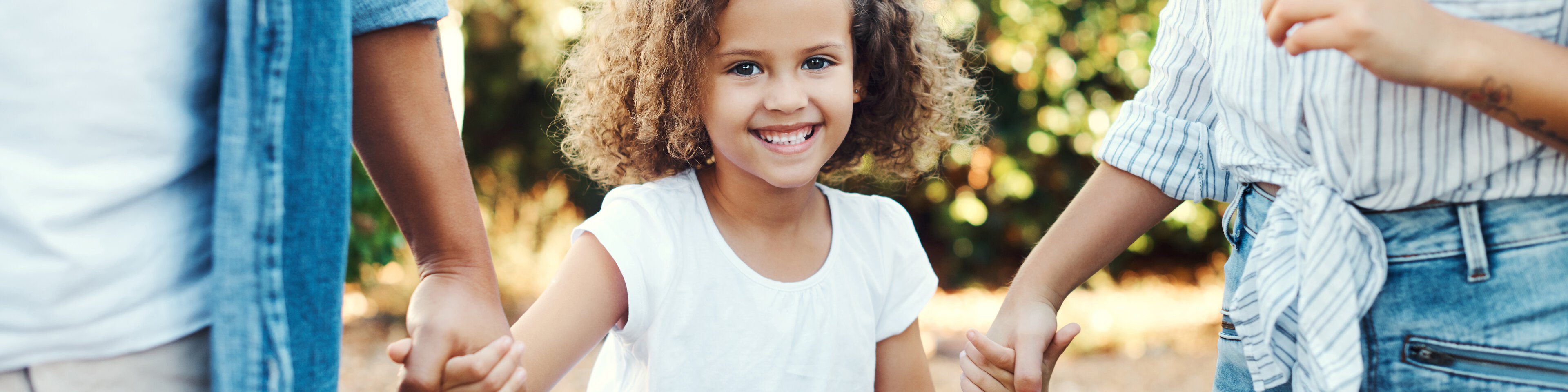 Ein kleines Mädchen mit braunen lockigen Haaren wird von seinen Eltern an beiden Händen gehalten | © PeopleImages - Getty Images/iStockphoto