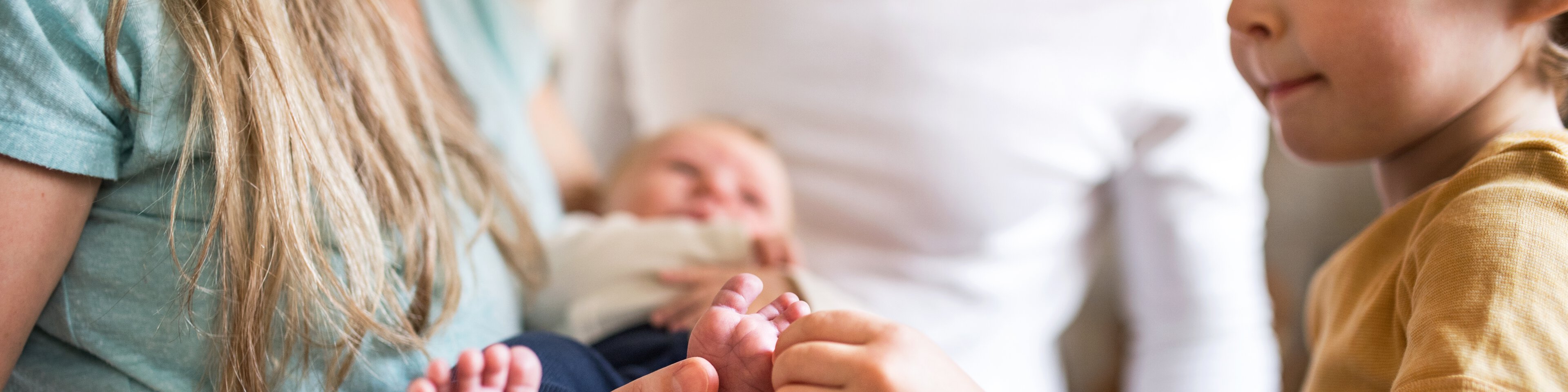 Eine Mutter hat ein Baby auf dem Arm und ein kleiner Junge drückt den kleinen Zeh des Babys | © Halfpoint - Getty Images/iStockphoto